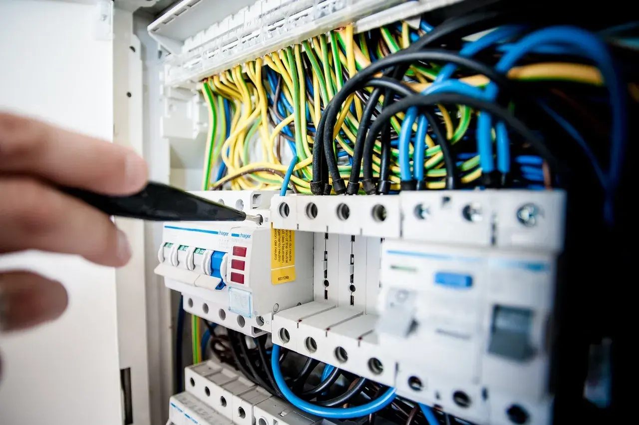 Elektryk naprawia awarię instalacji elektrycznej, manipulując śrubokrętem przy bezpiecznikach i plątaninie kolorowych przewodów.