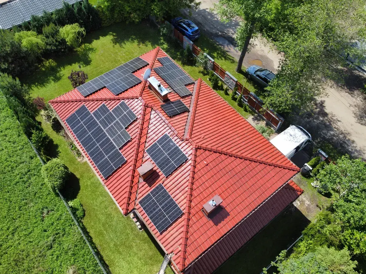 Panele fotowoltaiczne na czerwonym dachu, instalacja wykonana na starych zasadach. Wokół zieleń i samochody.