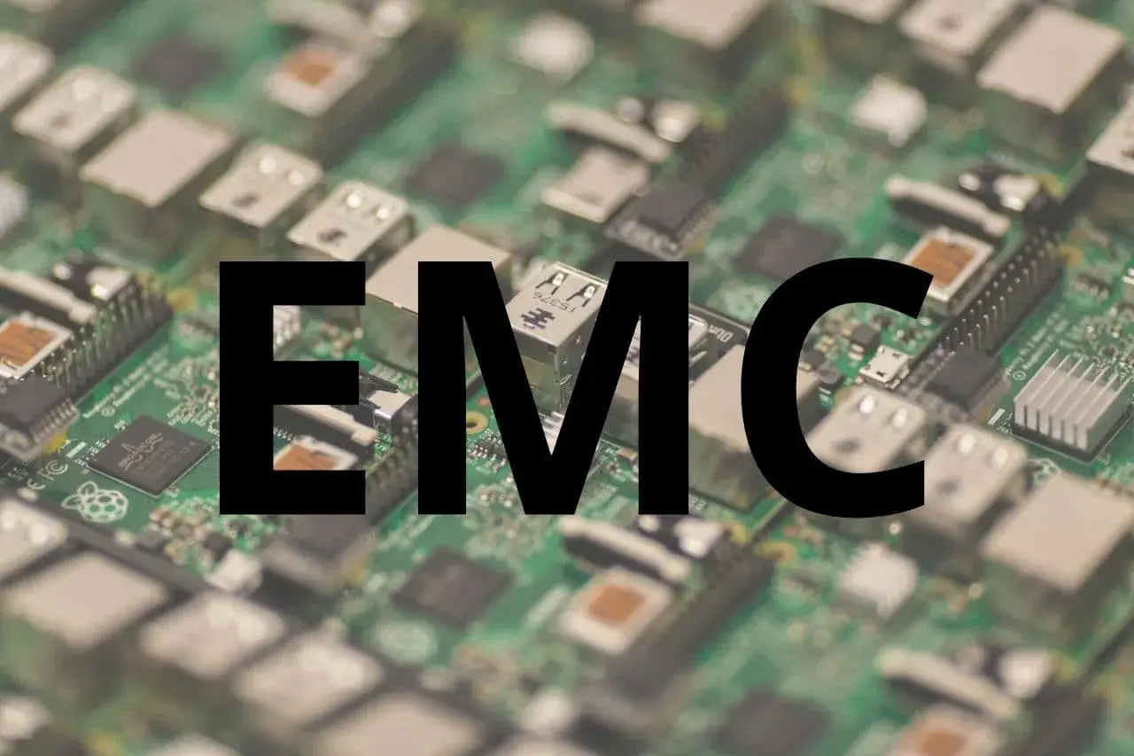 Na zielonej płytce drukowanej z widocznymi elementami elektronicznymi, wielkimi czarnymi literami widnieje napis "EMC". To skrót od Electromagnetic Compatibility, czyli kompatybilności elektromagnetycznej.