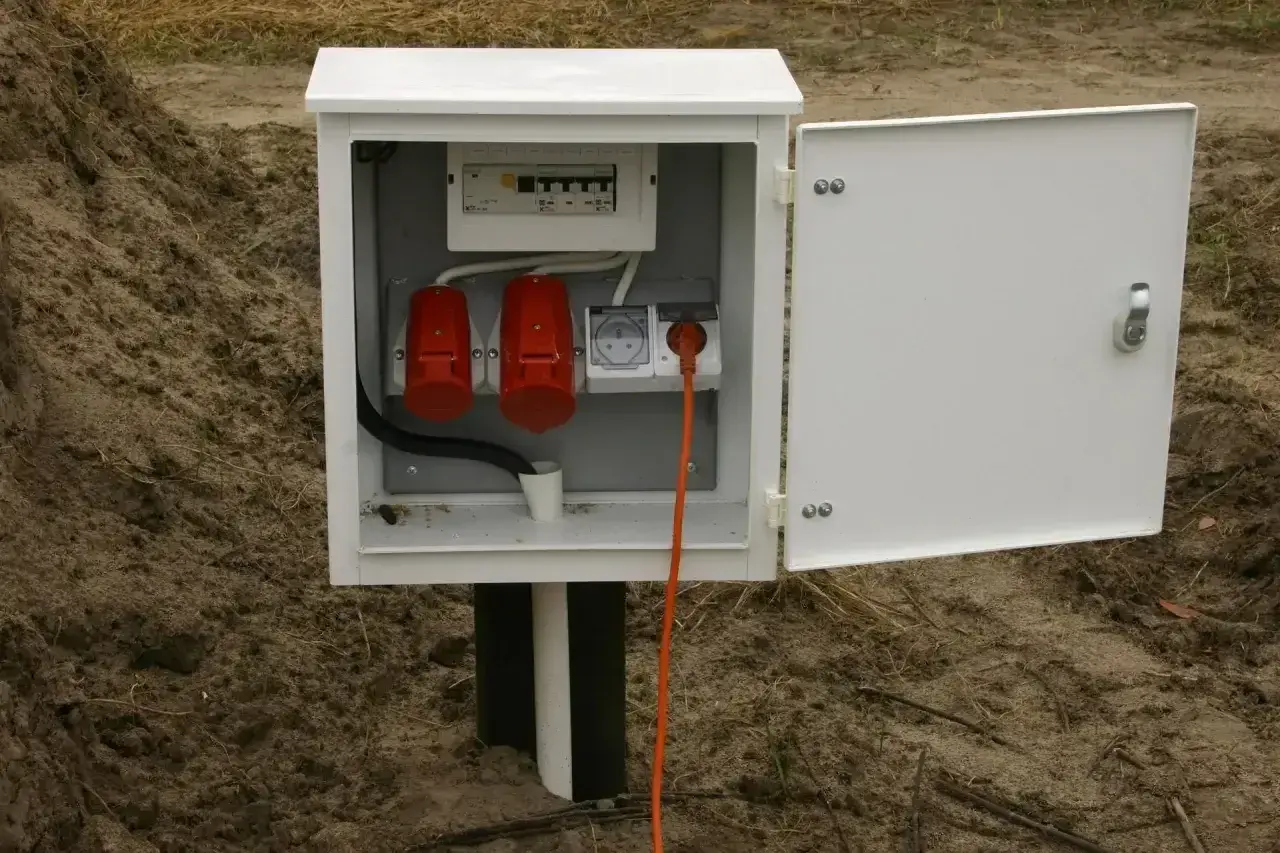 Biała skrzynka z bezpiecznikami i gniazdami, stanowiąca przyłącze elektryczne do domu. Pomarańczowy kabel wychodzi z gniazdka.