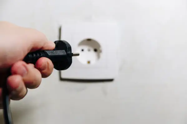Dłoń trzyma wtyczkę, zbliżając ją do gniazdka. Uważaj, by nie doświadczyć porażenie prądem z gniazdka!